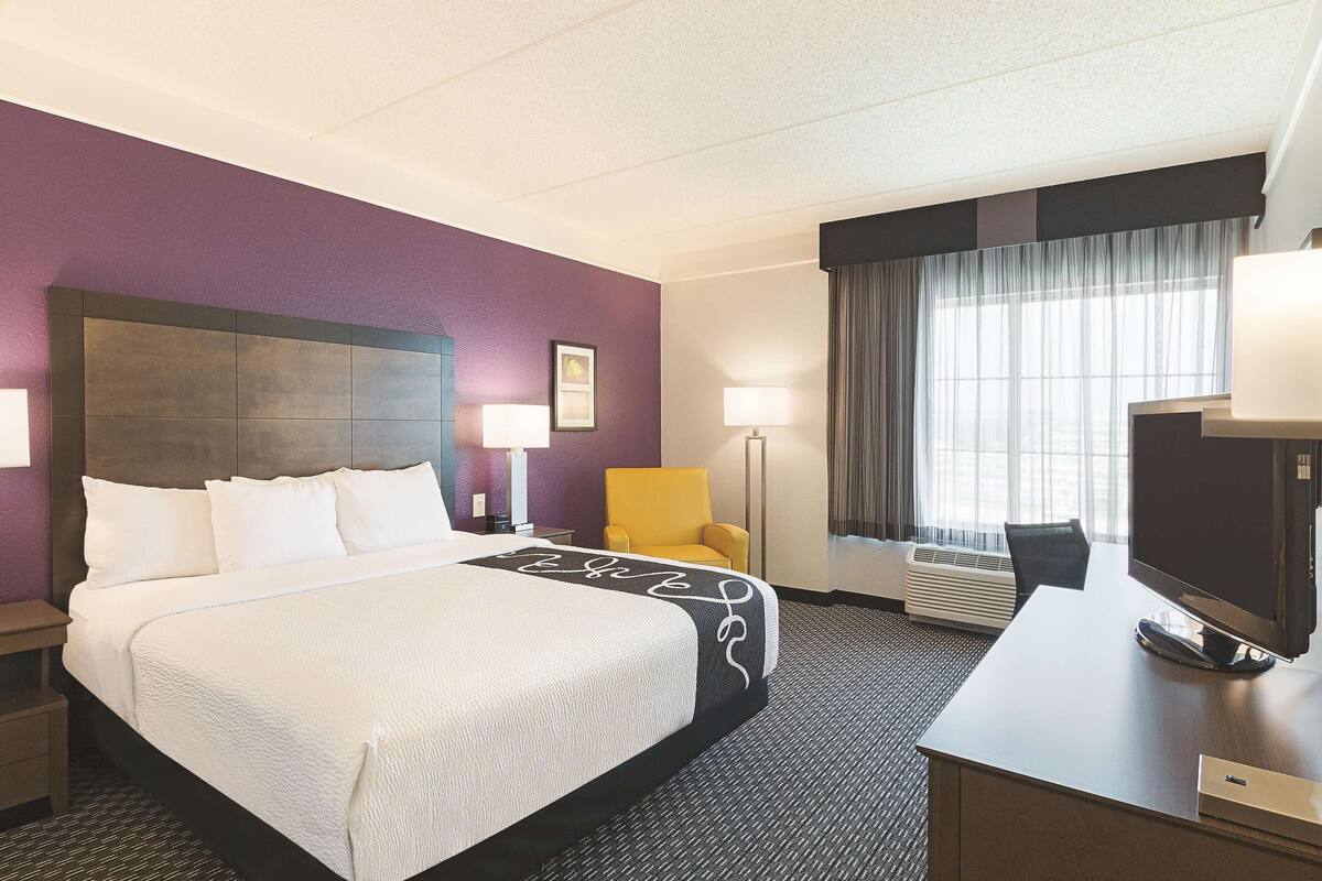 Una habitación de hotel con cama, escritorio, silla, televisor y ventana con cortinas.