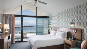 A hotel room with a large bed, a desk, a chair, a nightstand, and a view of the ocean.