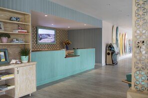 A reception area with a wooden reception desk, a television mounted on a patterned wall, and a row of surfboards against the wall.