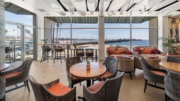 A spacious indoor area with wicker furniture, large windows offering a view of the beach and ocean, and a surf shop sign.
