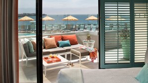 A balcony with a sofa, armchairs, and a table with drinks, overlooking a pool and umbrellas.