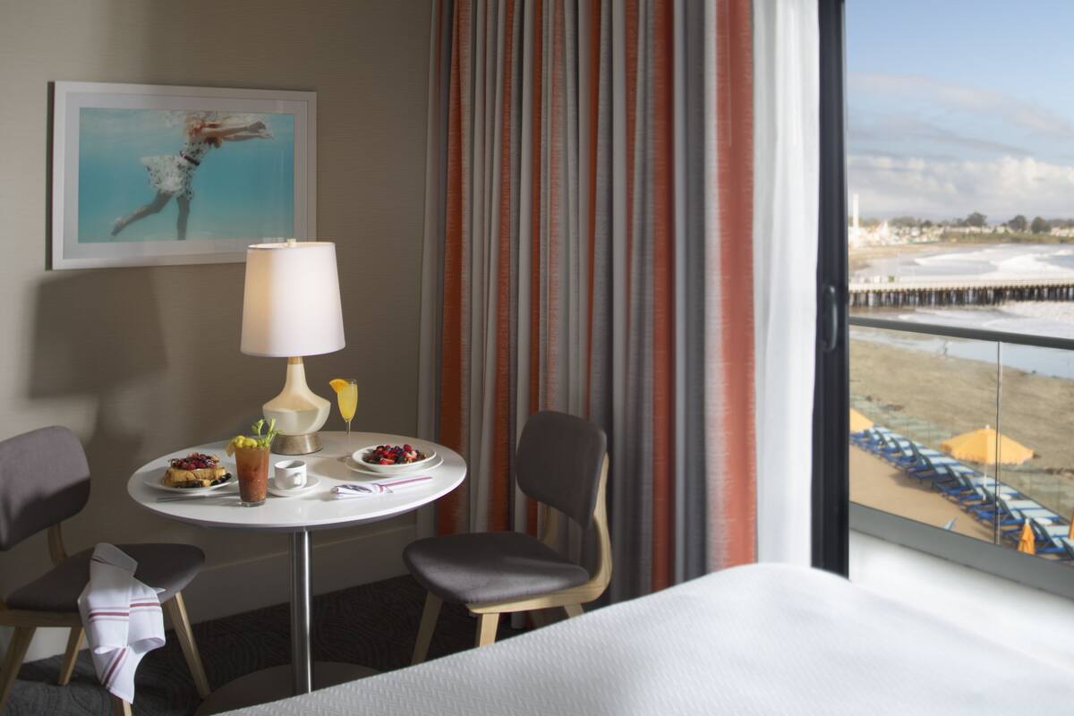 A hotel room with a bed, a dining table with a lamp, a chair, a small table with a drink and fruit, and a view of the beach through the window.