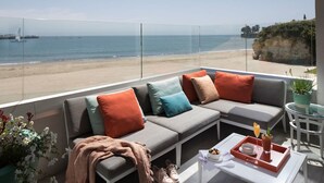 A balcony with a view of the beach, a sofa with cushions, a glass table with a tray of food and drinks, and a potted plant.