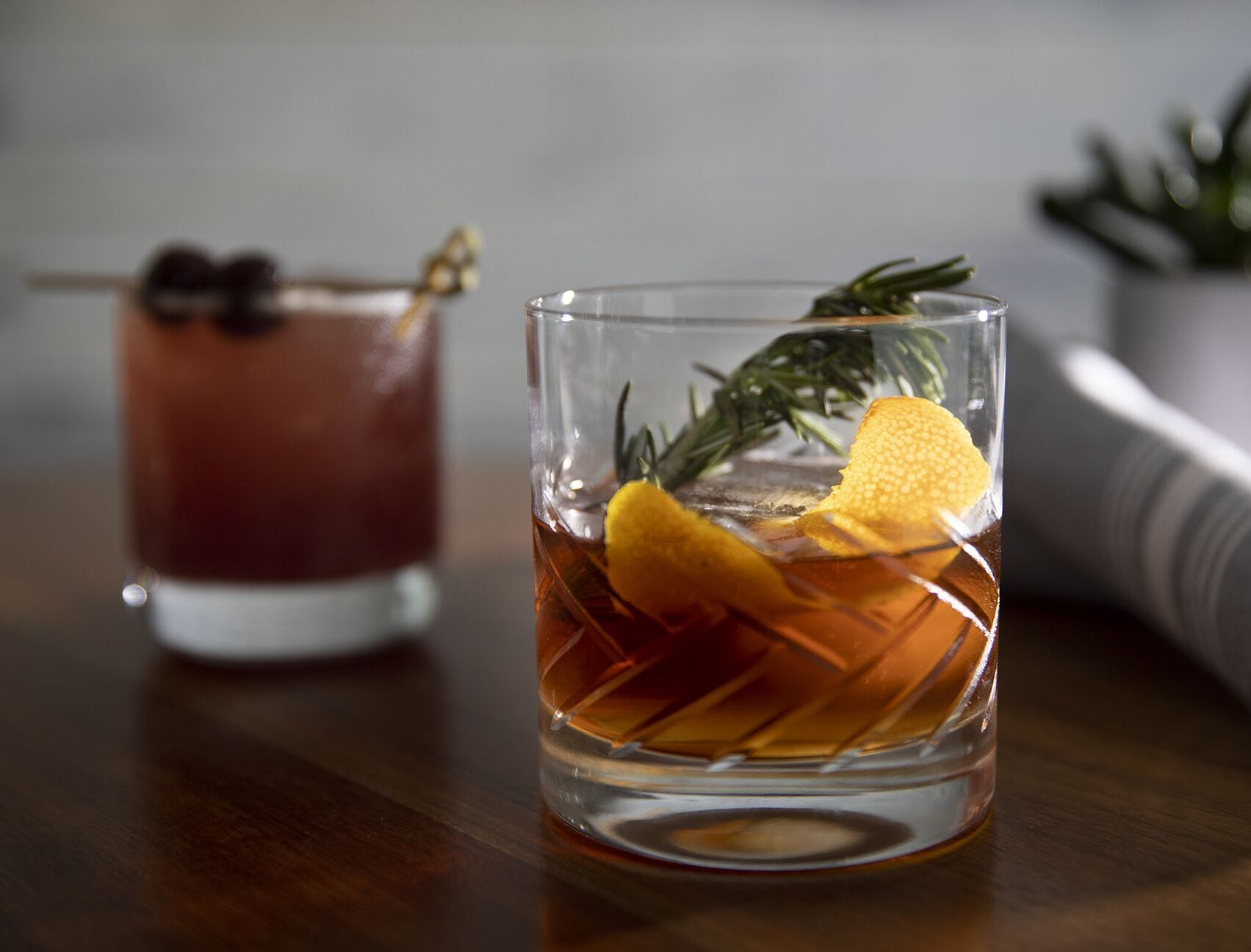 Two glasses of amber-colored beverages with lemon slices and rosemary, placed on a wooden surface.
