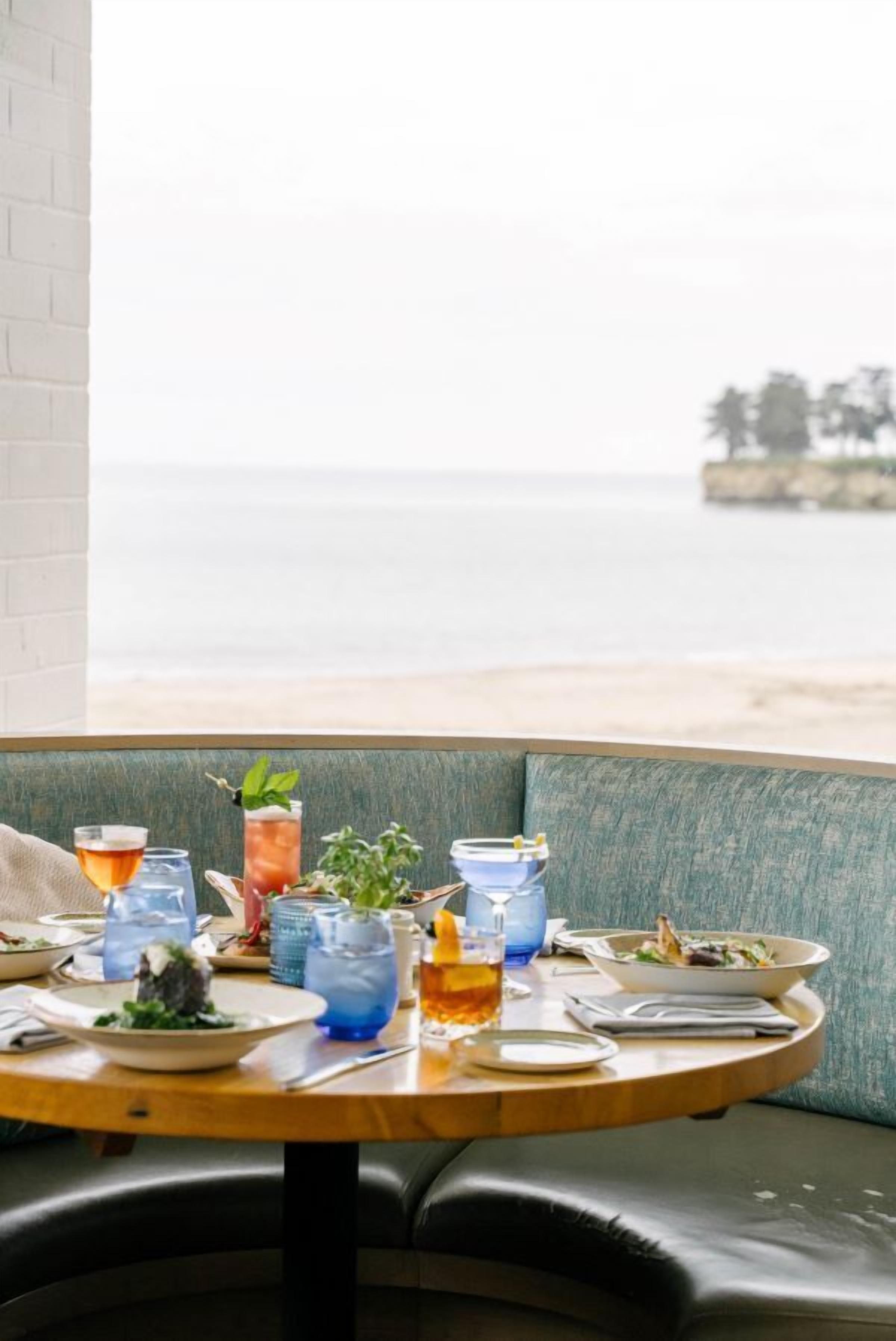 A table set with drinks and plates of food, overlooking a beach.