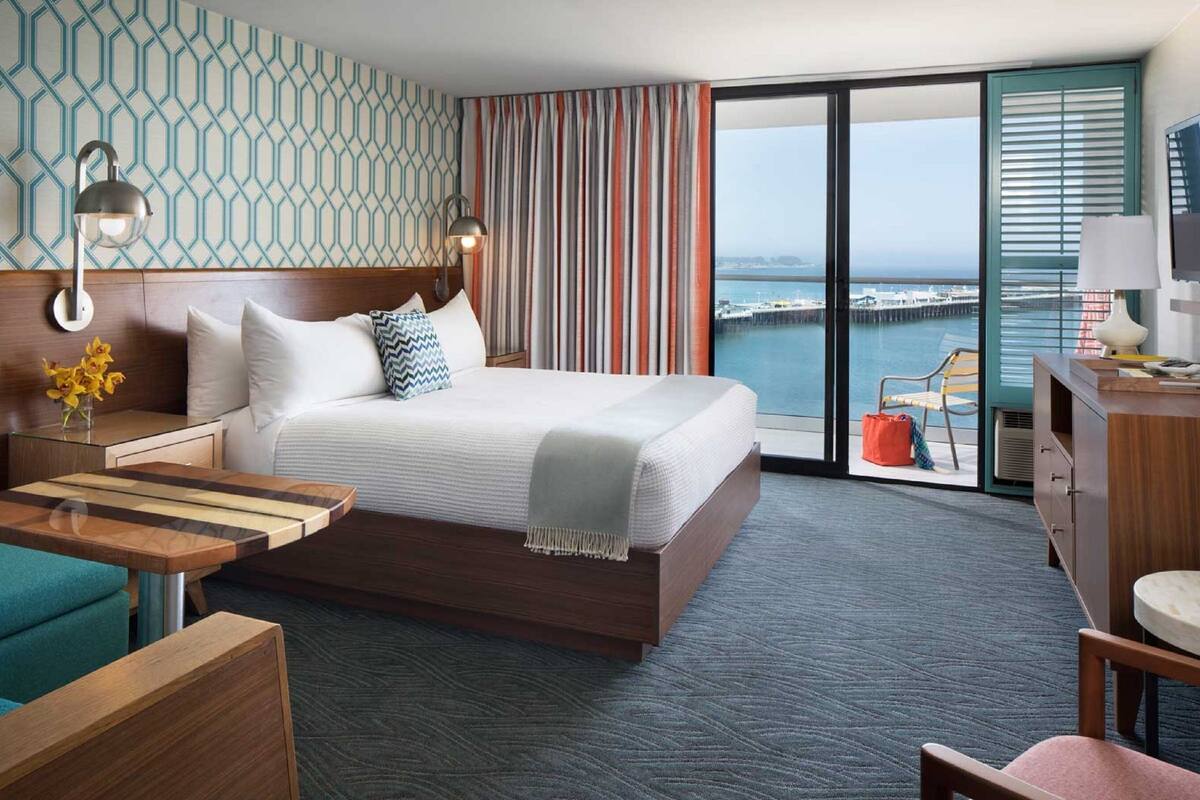 A modern hotel room with a large bed, a view of the sea, a balcony with a chair, and a desk with a lamp.