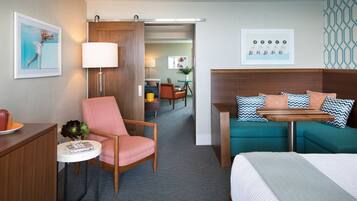 A hotel room with a bed, a sofa, a chair, a small table, and a desk. There is a lamp, a picture frame, and a plant on the desk.