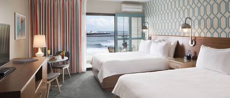 A hotel room with two beds, a desk, a chair, a small table with a lamp, and a view of the sea.