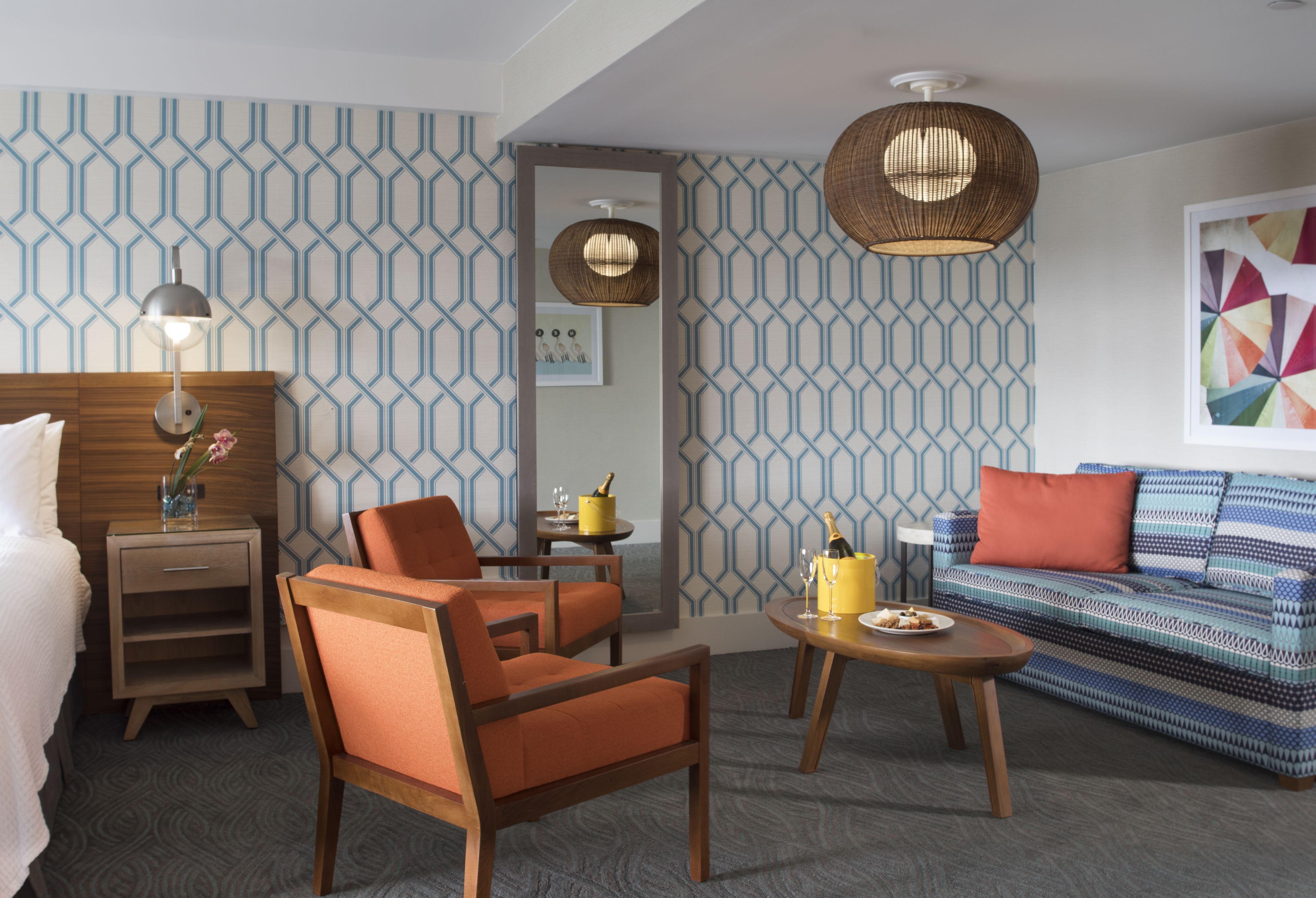 A hotel room with a patterned wallpaper, a wooden headboard, a sofa, two armchairs, a coffee table, a side table with a lamp, and a framed picture on the wall.