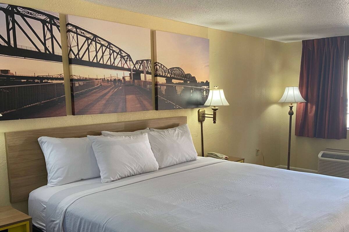 Una habitación de hotel con una cama grande, dos lámparas fijadas a la pared, un teléfono y un cuadro de un puente.