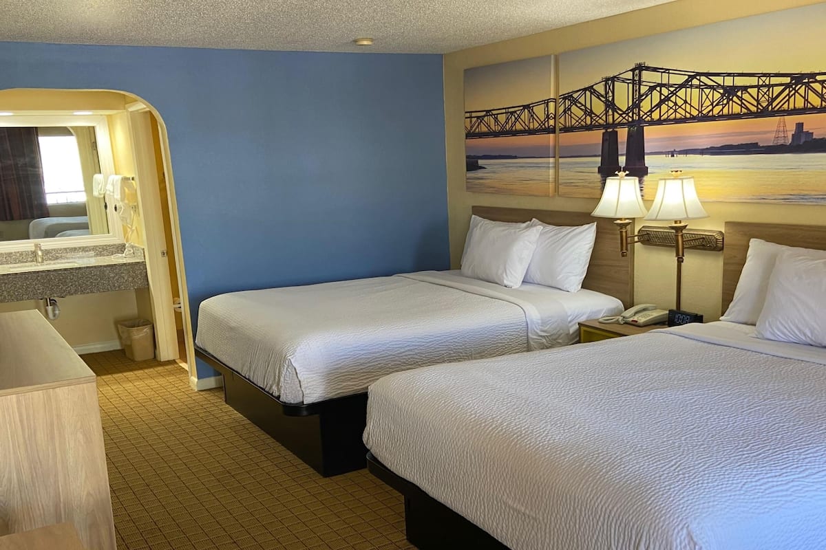 Habitación de hotel con dos camas, un baño con espejo y un cuadro de un puente.