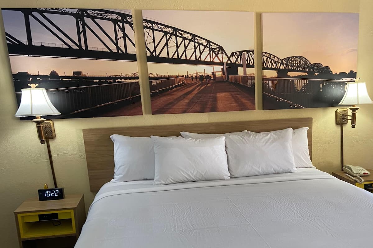 Habitación de hotel con una cama, dos lámparas de noche, una mesita de noche con un reloj digital y tres cuadros con imágenes de un puente.