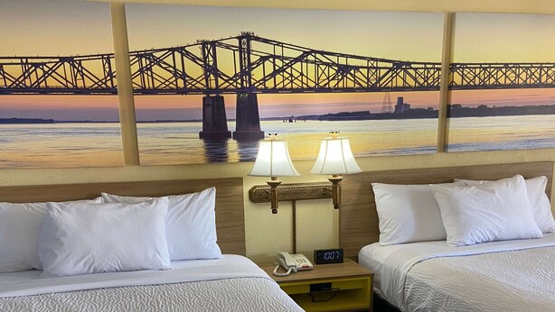 Ein Hotelzimmer mit zwei Betten, weißer Bettwäsche, einem Nachttisch mit Telefon und Lampe sowie einem Bild einer Brücke an der Wand.