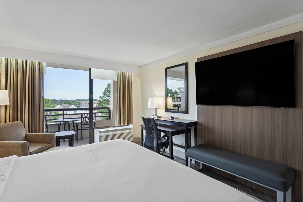 Quarto de hotel com uma cama grande, uma mesa com cadeira, um banco, uma TV e uma varanda com cortinas.