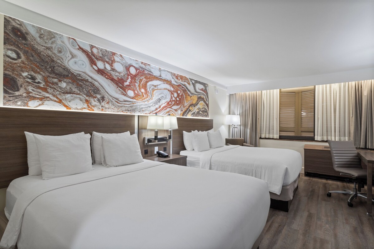 Quarto de hotel com duas camas, uma escrivaninha, uma cadeira, um quadro abstrato grande na parede e uma janela com cortinas.