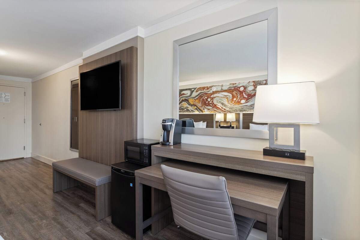 Quarto de hotel com secretária de madeira, televisão de tela plana na parede, cafeteira, luminária e um banco.