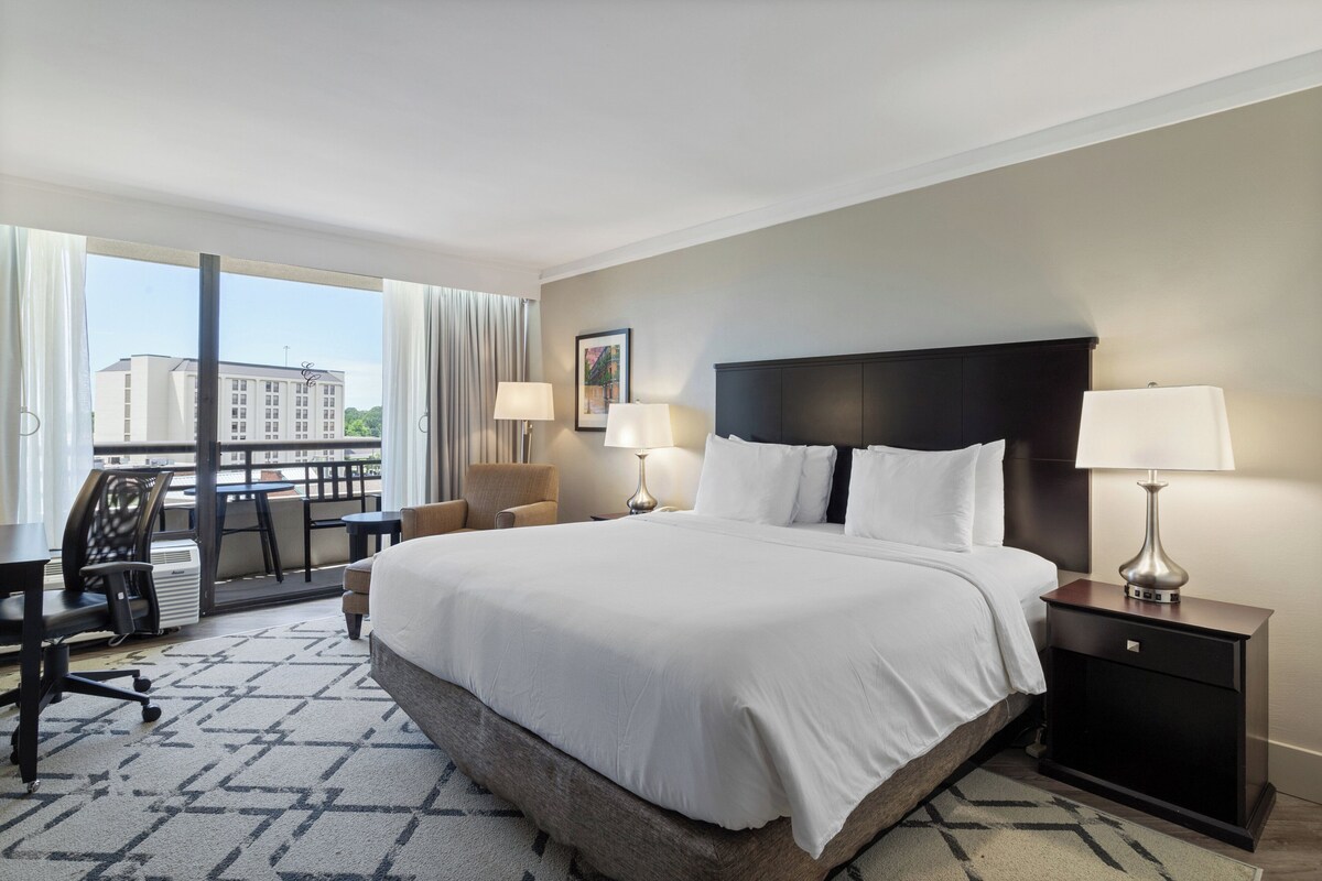 Quarto de hotel com uma cama grande, duas mesas de cabeceira com abajures, uma escrivaninha com cadeira e vista para um edifício pela janela.