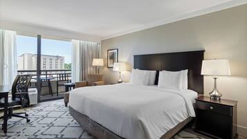A hotel room with a large bed, two bedside tables with lamps, a desk with a chair, and a view of a building through the window.