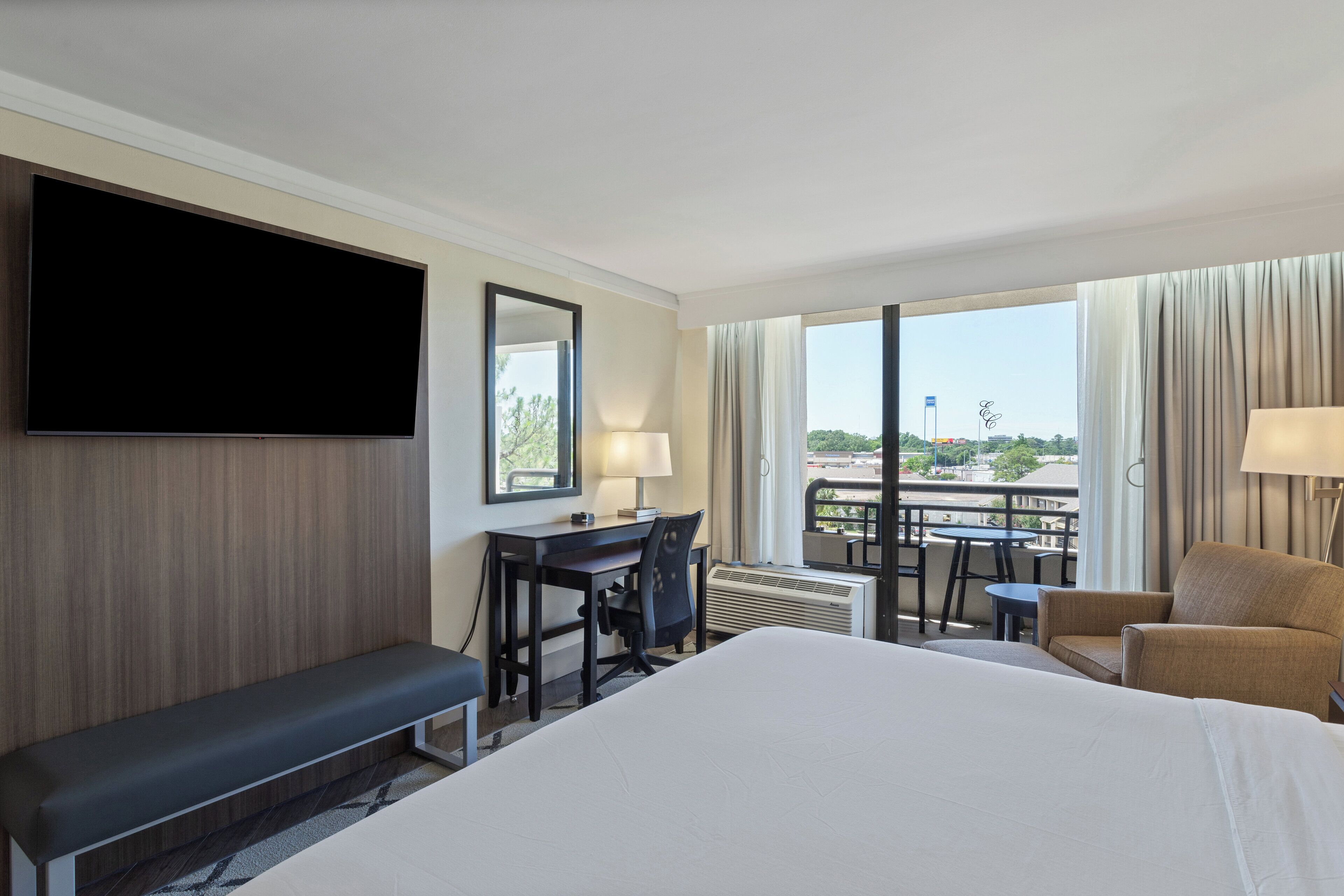 Habitación de hotel con una cama grande, un escritorio con silla, televisión montada en la pared y un balcón con vistas a la ciudad.