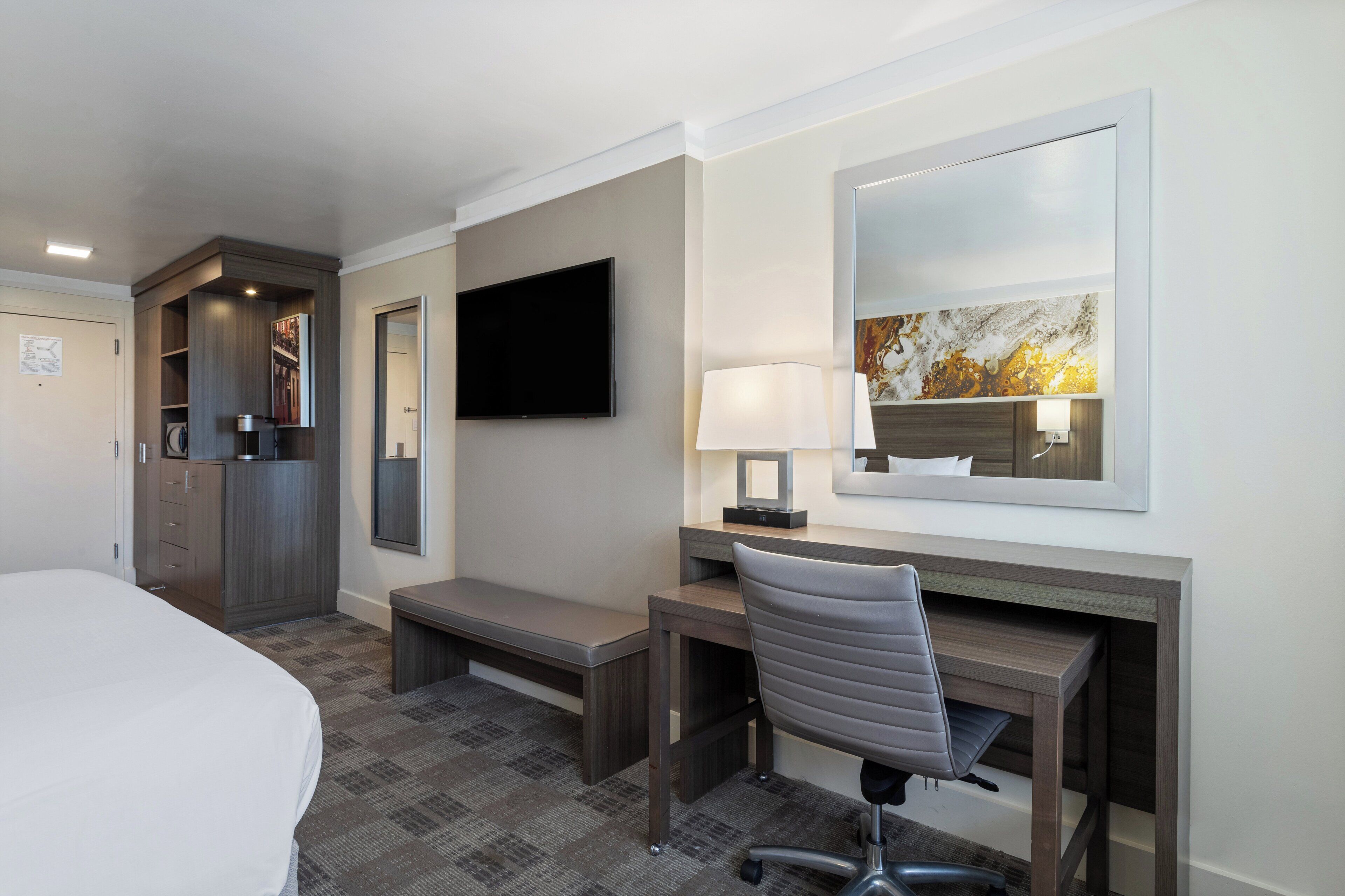 A hotel room with a bed, a desk with a chair, a television, a lamp, and a painting on the wall.