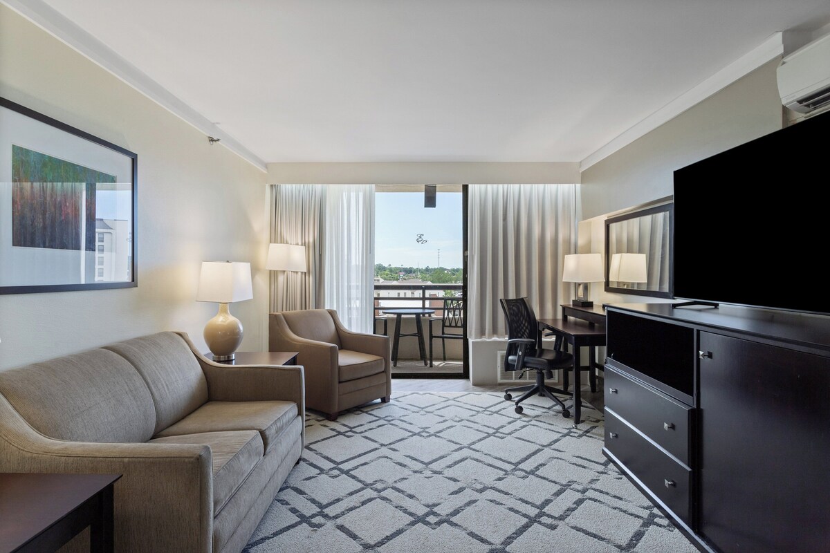 Quarto de hotel com sofá, duas cadeiras, escrivaninha, televisão e varanda com cortinas.