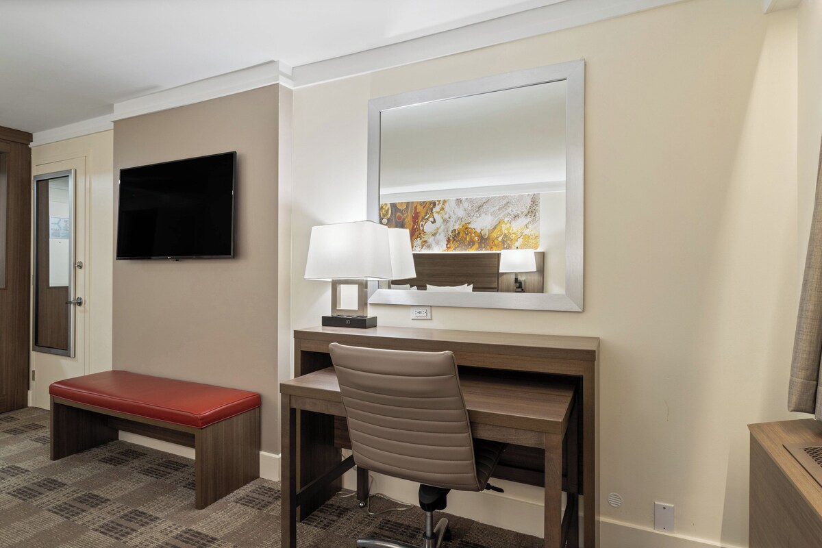 Quarto de hotel com escrivaninha, cadeira, televisão e um banco.