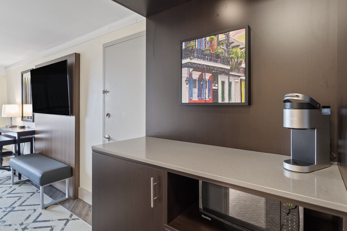 Quarto de hotel moderno com cozinha compacta, televisão de tela plana, cafeteira e pintura de uma cena urbana.