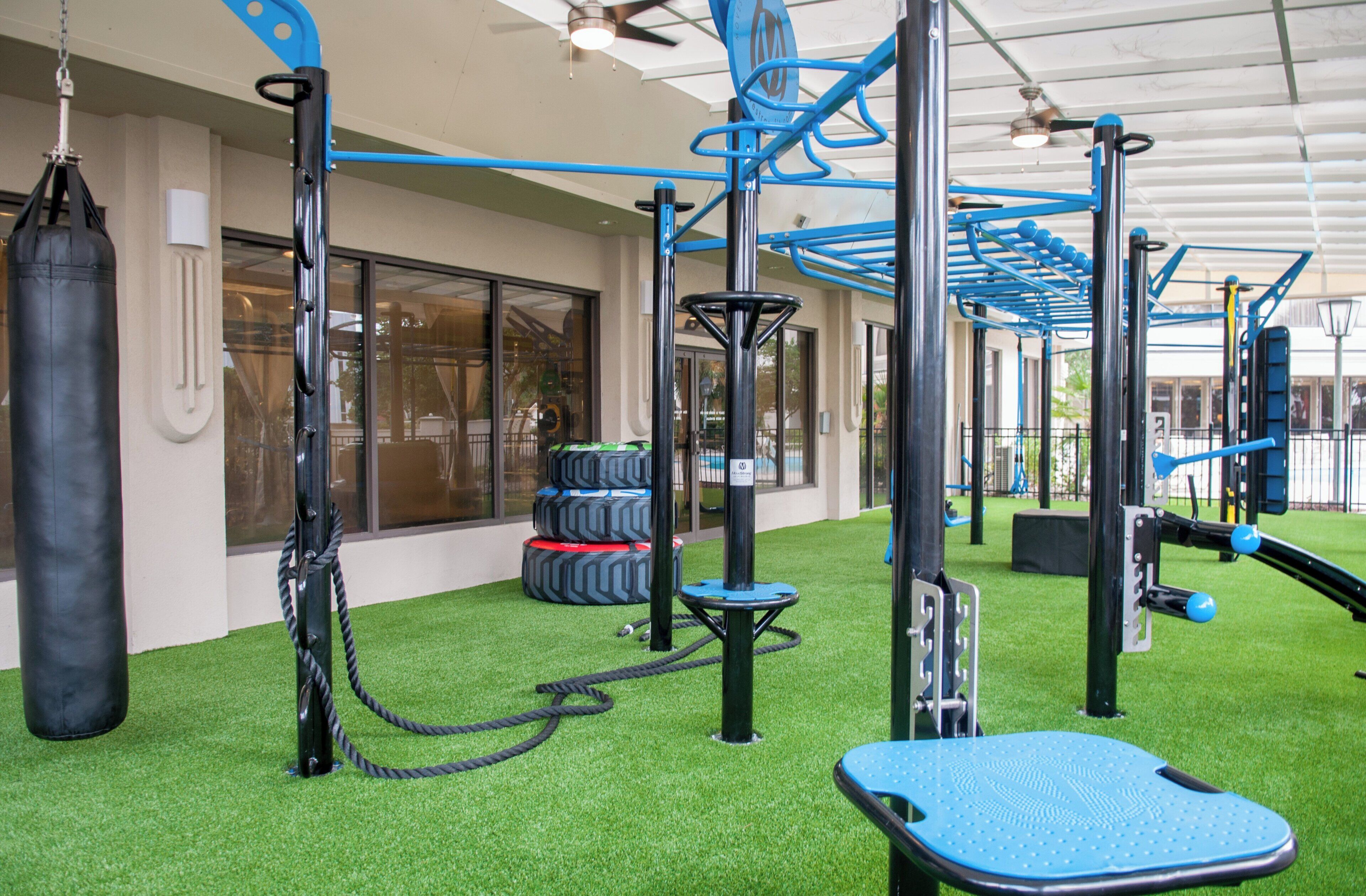 An outdoor gym area with exercise equipment, artificial turf, and a building with large windows in the background.