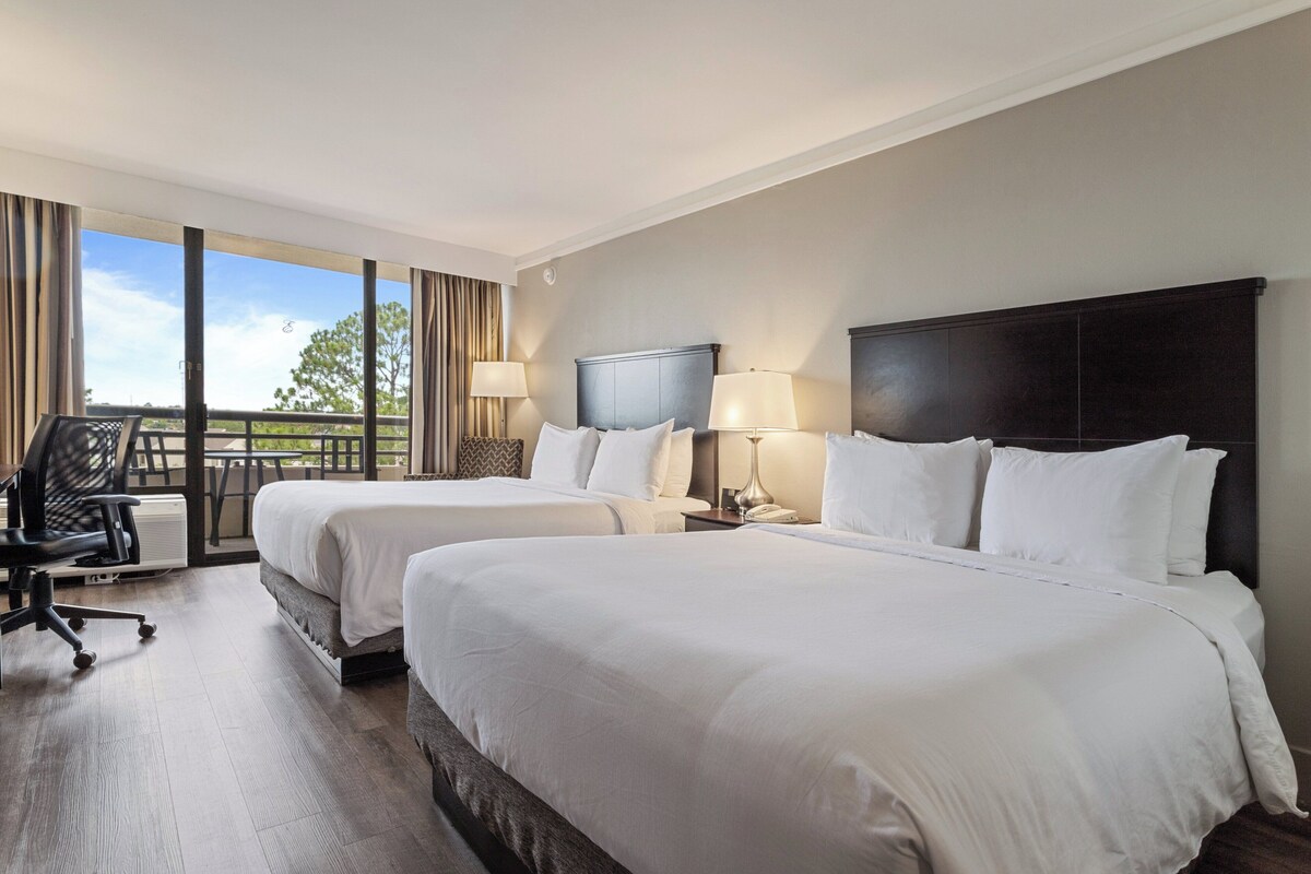 Quarto de hotel com duas camas, uma escrivaninha, uma cadeira e uma grande janela com vista para o exterior.