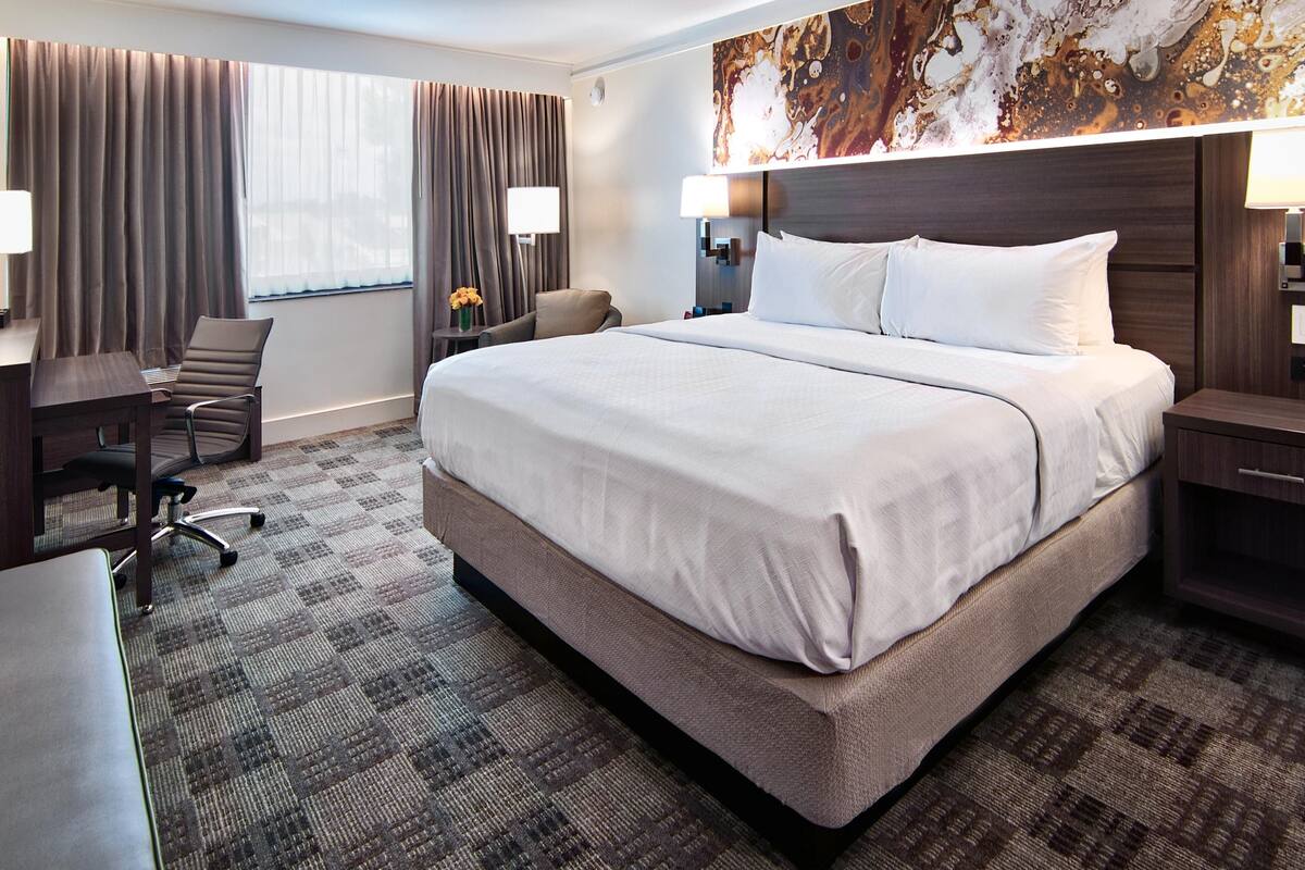 A hotel room with a large bed, a desk with a chair, a small table with a lamp, and a large abstract painting on the wall.