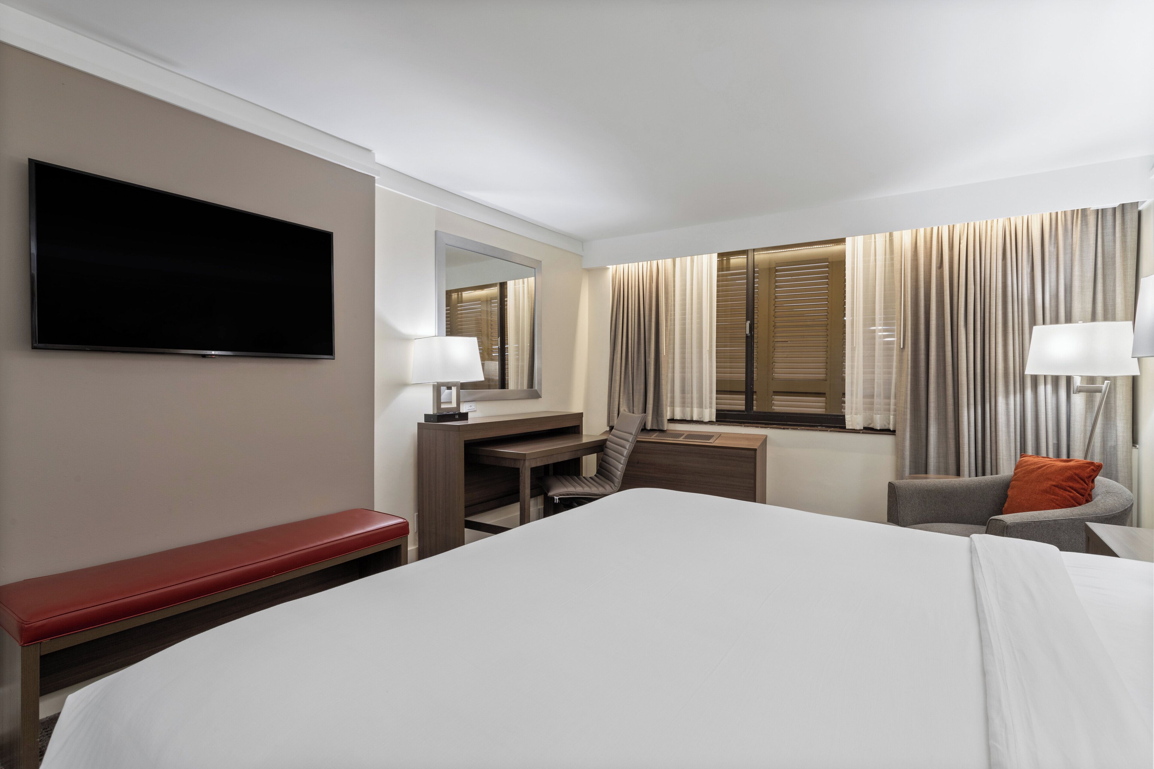 Quarto de hotel com uma cama grande, uma escrivaninha, uma cadeira, uma televisão e uma janela com cortinas.