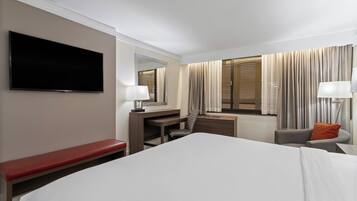 Quarto de hotel com uma cama grande, uma escrivaninha, uma cadeira, uma televisão e uma janela com cortinas.