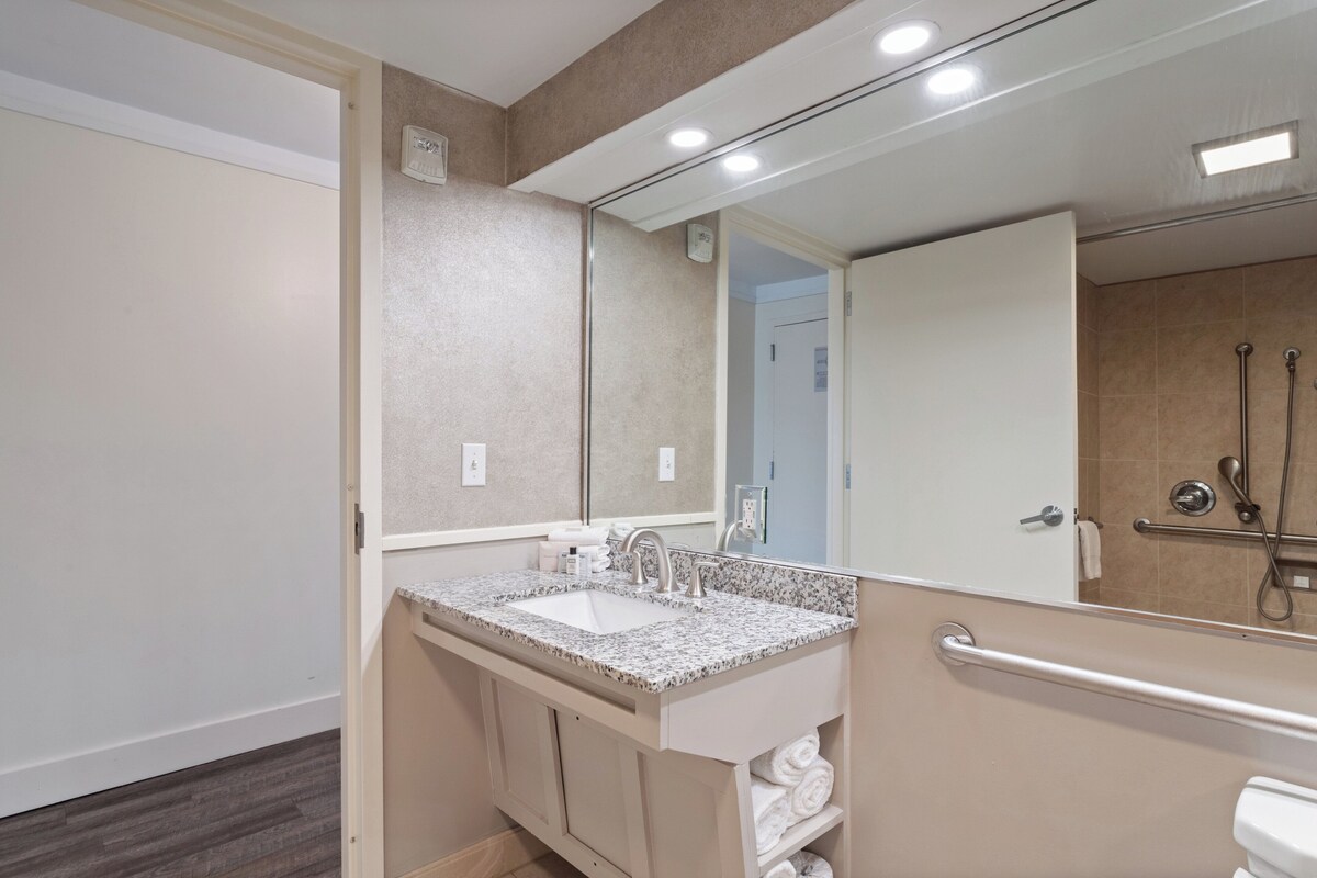Uma casa de banho moderna com lavatório bancada de granito, um espelho grande e um chuveiro amplo com barra de apoio.