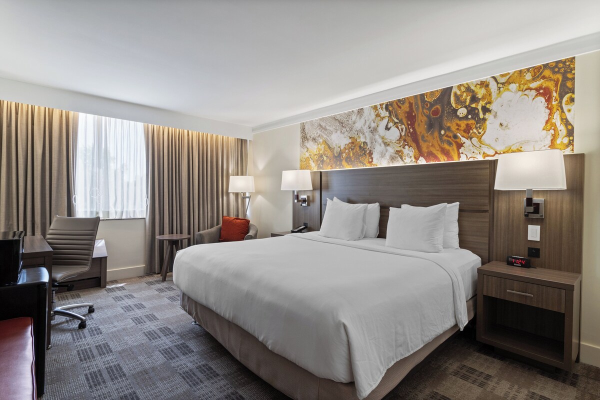 Quarto de hotel com uma cama grande, duas mesas de cabeceira com luminárias, uma escrivaninha com cadeira e um quadro abstrato grande na parede.