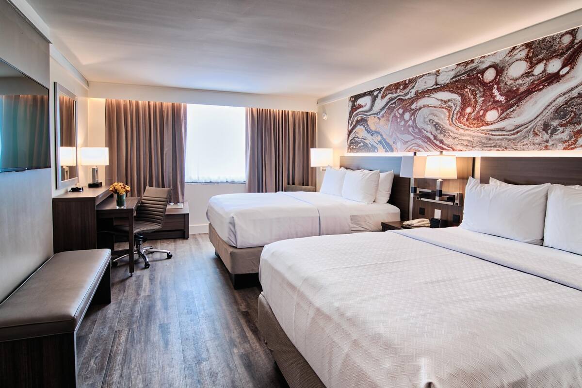 A hotel room with two beds, a desk, a chair, a bench, and a large abstract painting on the wall.