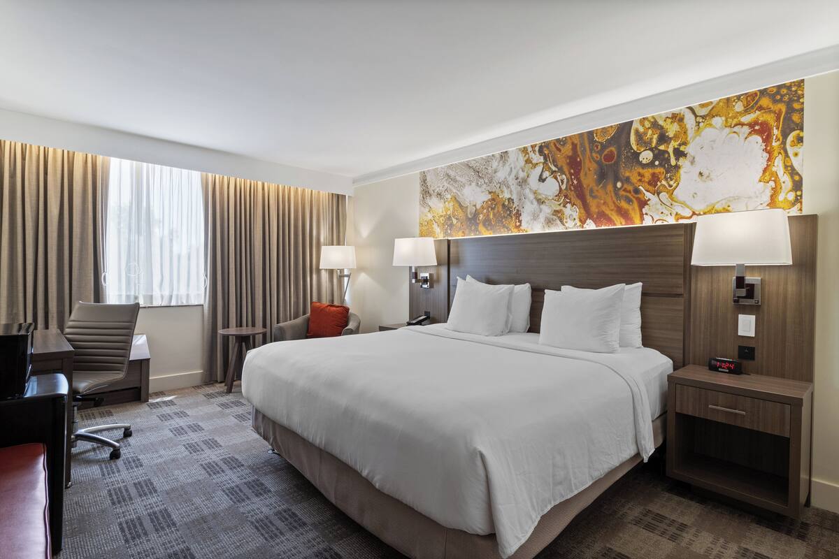 A hotel room with a large bed, two bedside tables with lamps, a desk with a chair, and a large abstract painting on the wall.