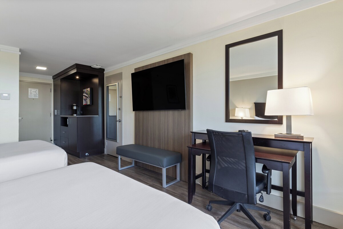 Quarto de hotel com duas camas, uma escrivaninha com cadeira, televisão, espelho e abajur.