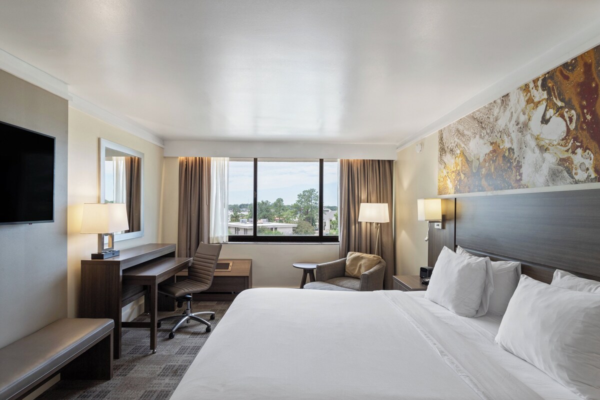 Quarto de hotel com cama grande, escrivaninha, cadeira, televisão e janela com cortinas.