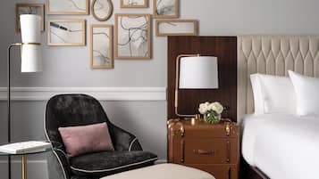 Habitación de hotel con cama, una silla, una mesita, lámpara, cómoda y obras de arte en la pared.
