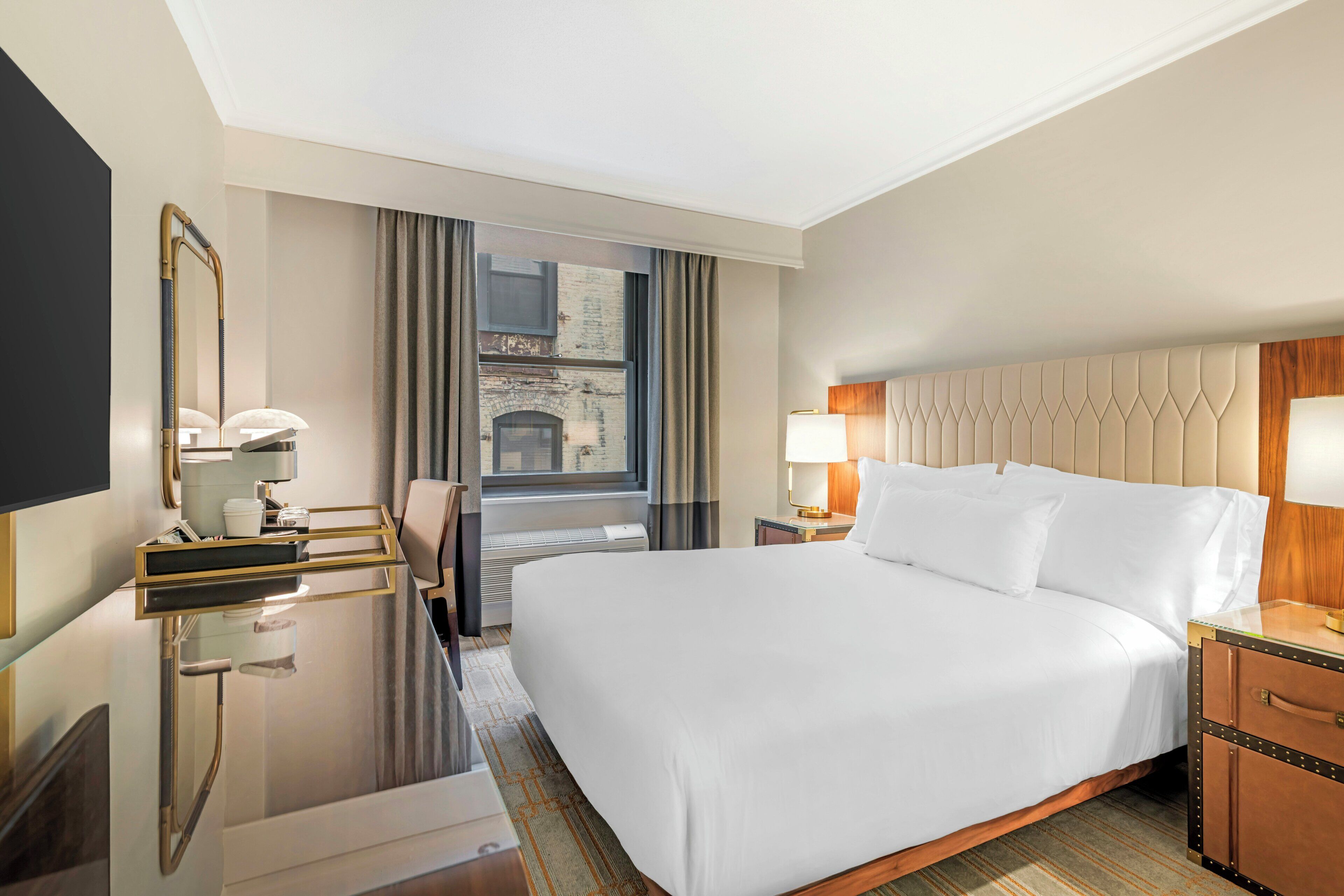 Ein Hotelzimmer mit einem großen Bett, einem Schreibtisch mit Spiegel, einem Fenster mit Vorhängen und Blick auf ein Gebäude.