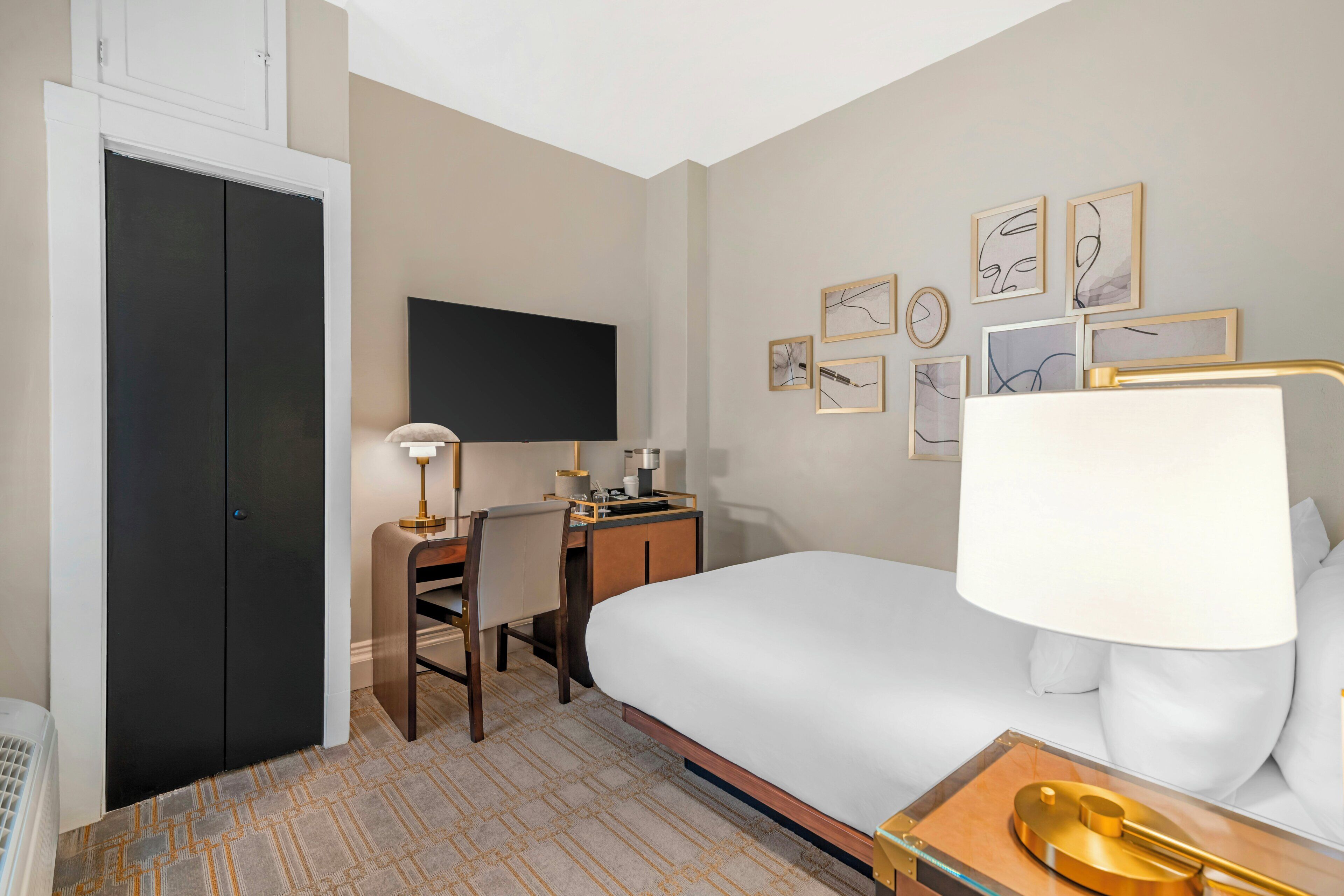 A hotel room with a bed, desk, chair, flat-screen TV, and artwork on the wall.