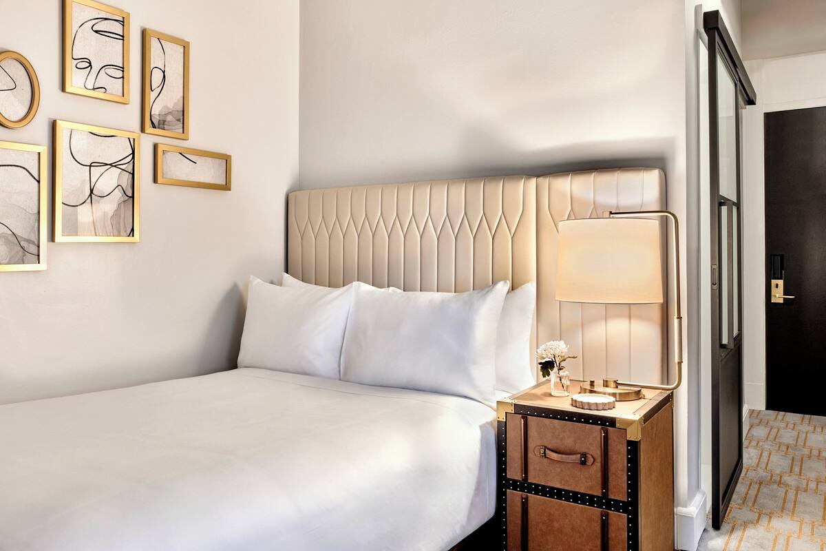 Une chambre d’hôtel avec un lit, une table de chevet avec une lampe, une petite armoire et une œuvre d’art abstraite au mur.