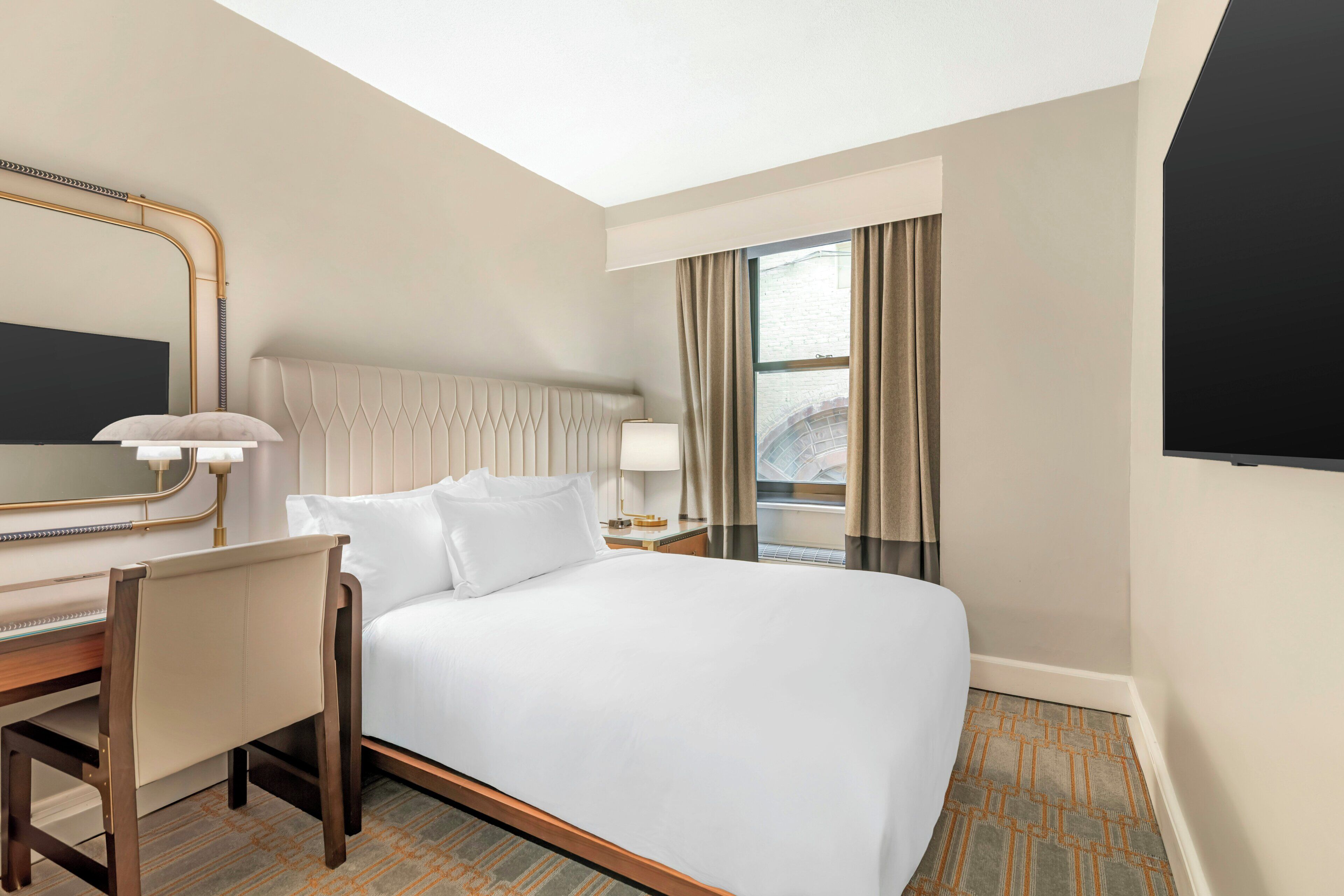Une chambre d’hôtel équipée d’un lit, d’un bureau, d’une chaise, d’un miroir, d’une fenêtre avec des rideaux et d’un téléviseur à écran plat.