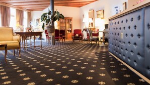 Una habitación con alfombra estampada, muebles de madera, un aparador tapizado azul y una planta grande.