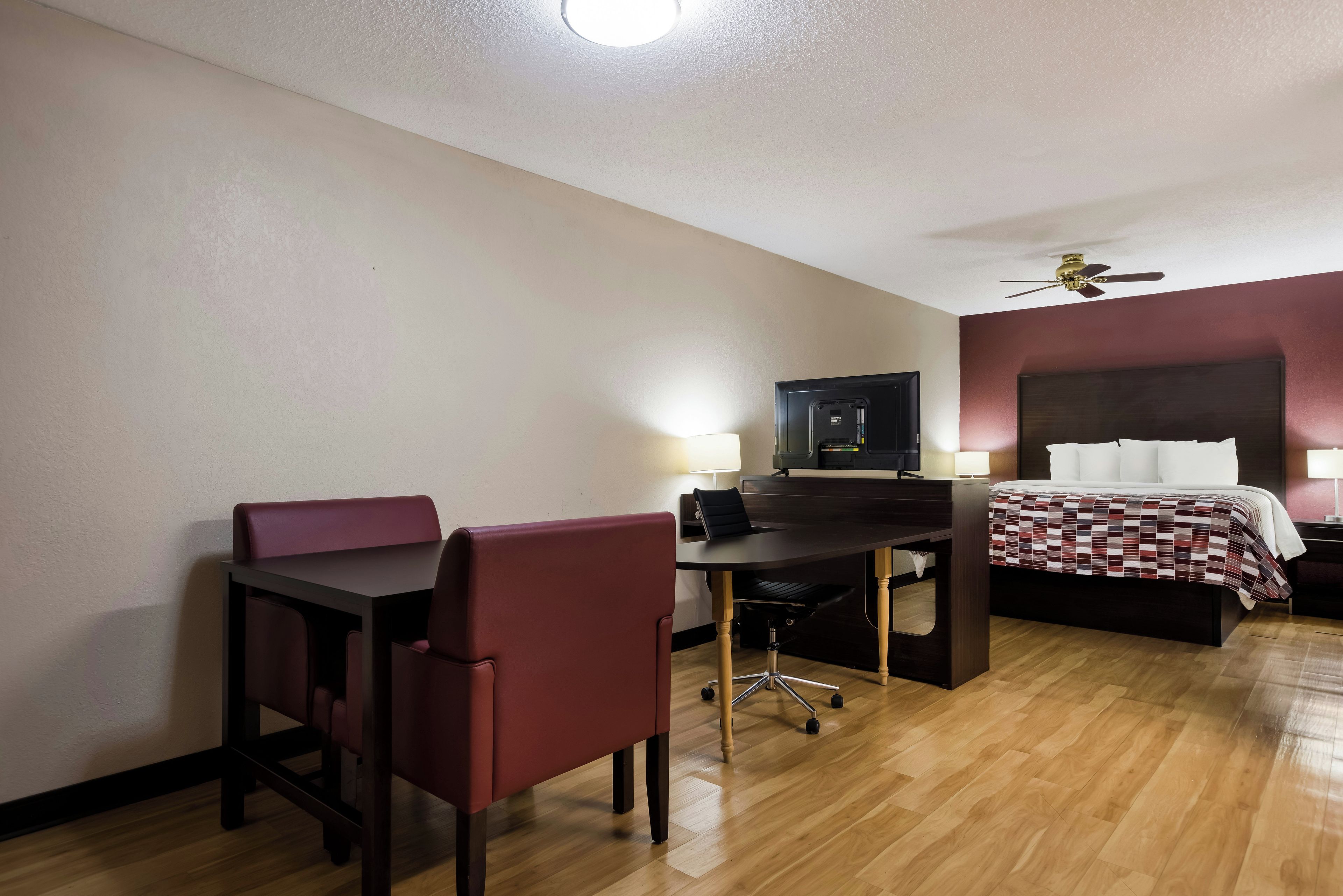 A hotel room with a desk, two chairs, a bed, a ceiling fan, and a television.