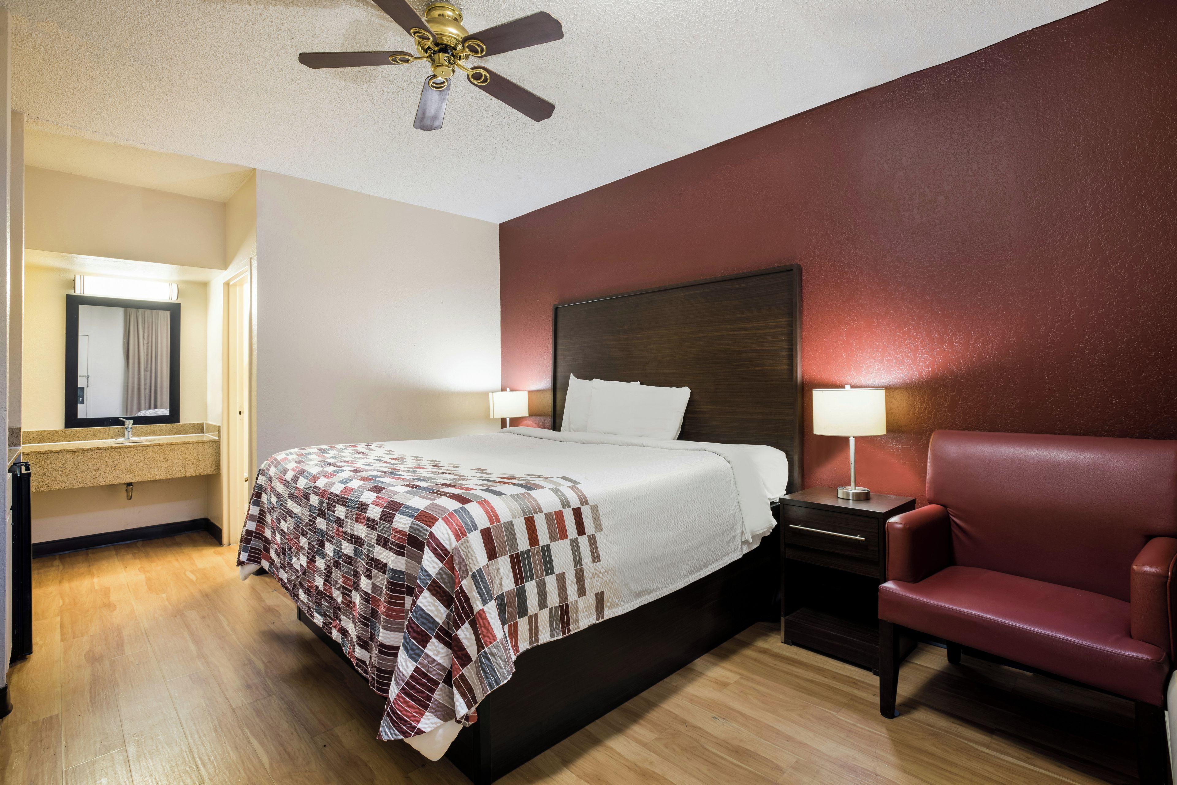 A hotel room with a bed, a chair, a nightstand, a lamp, a ceiling fan, and a mirror.