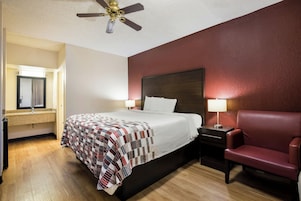 A hotel room with a bed, a chair, a nightstand, a lamp, a ceiling fan, and a mirror.