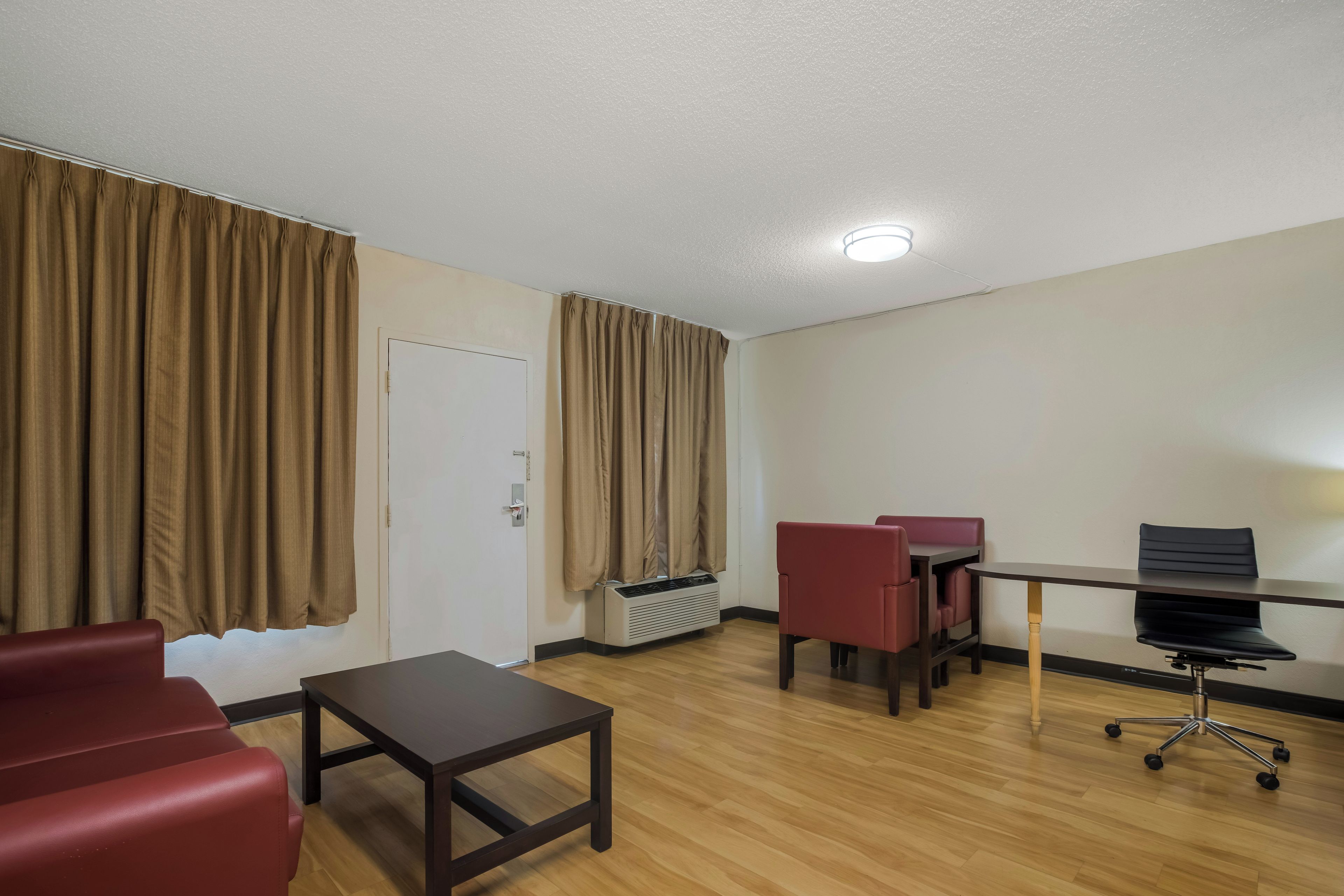 A hotel room with a wooden floor, a desk with a chair, a red sofa, and a door with curtains.