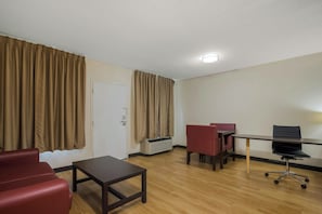 A hotel room with a wooden floor, a desk with a chair, a red sofa, and a door with curtains.