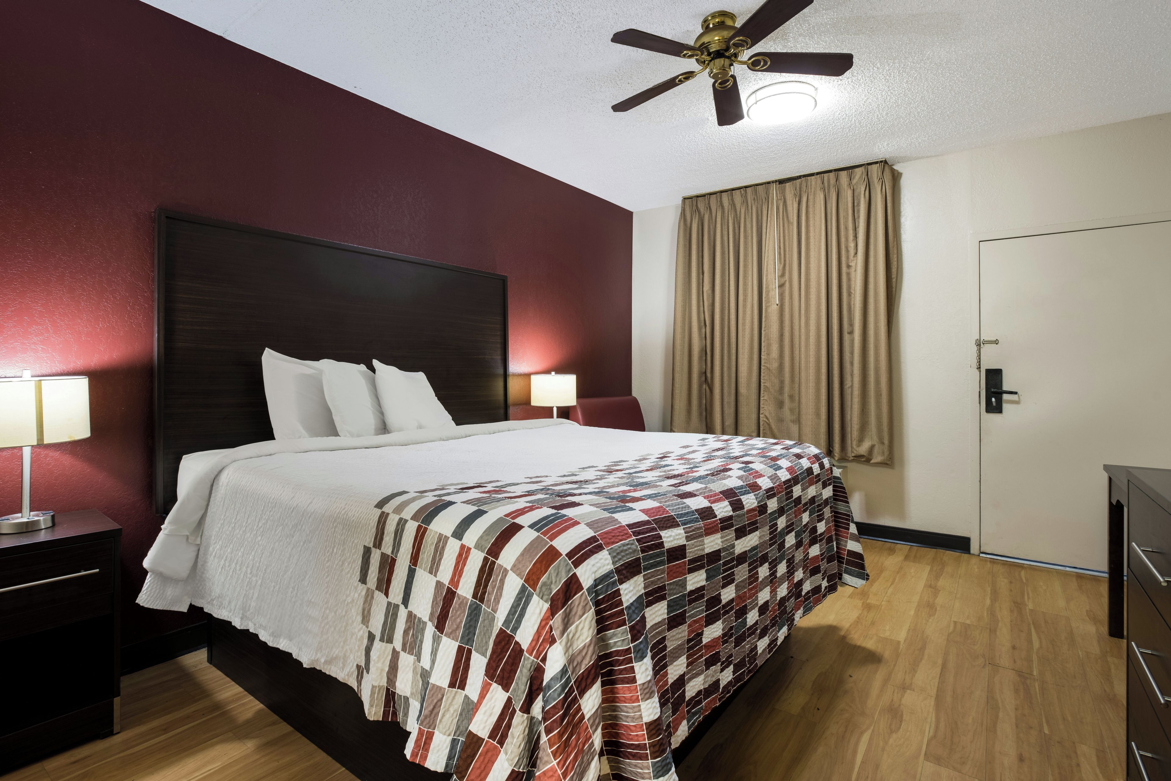 A hotel room with a large bed, two bedside lamps, a ceiling fan, a window with curtains, and a door.